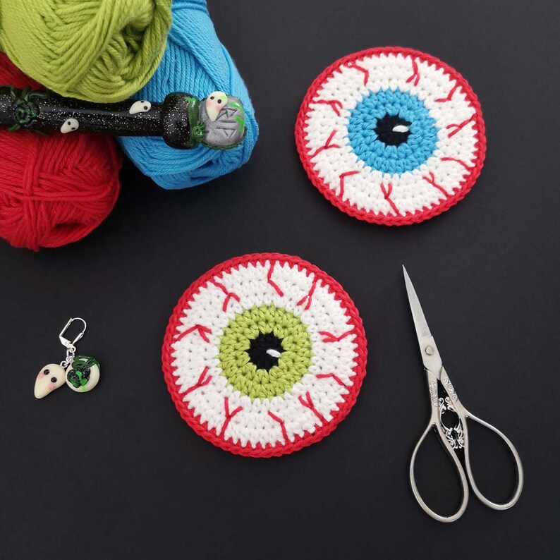 May include: Two crocheted eye-shaped coasters made with red, white and green yarn. The coasters are on a black surface with a pair of silver scissors.