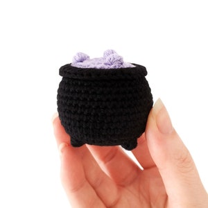 May include: A small black crocheted cauldron with a purple crocheted lid. The cauldron is being held in a hand.