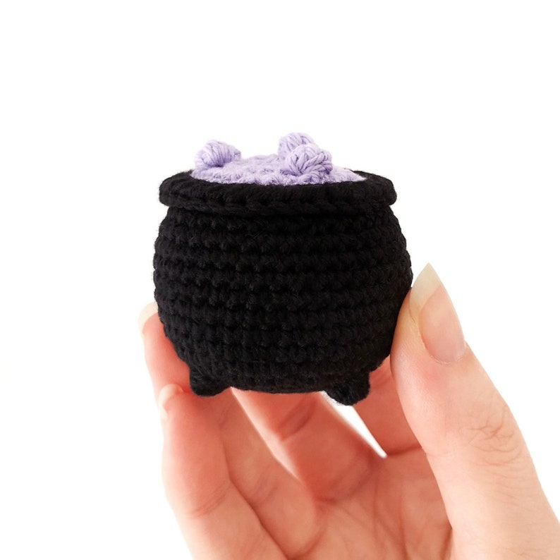 May include: A small black crocheted cauldron with a purple crocheted lid. The cauldron is being held in a hand.