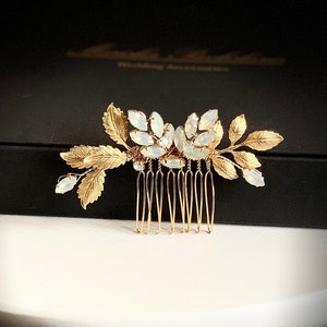 May include: Gold hair comb with a floral design and white opal stones. The comb is on a black box with the text "Wedding Accessories".