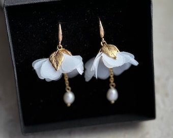 May include: A pair of white flower earrings with gold accents and pearl drops. The earrings are displayed in a black box.