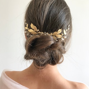 May include: A gold hair comb with leaf and crystal accents. The comb is worn in a woman's hair, which is styled in a low bun.