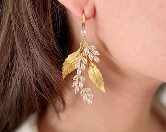 Puede incluir: Pendientes colgantes de oro con un diseño de hojas y detalles de cristal transparente.