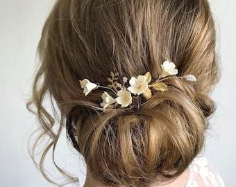 Puede incluir: Un peine de pelo floral blanco con detalles dorados. El peine está hecho de pequeñas flores blancas y hojas doradas. Es un accesorio para el cabello delicado y elegante.