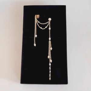 Könnte beinhalten: Ein goldfarbener Ohrschmuck mit herabhängenden Ketten und Kunstperlen. Der Ohrschmuck ist mit einer Reihe von filigranen Ketten verbunden, die nach unten hängen und mit kleinen, runden, weißen Perlen verziert sind. Der Schmuck wird auf einer schwarzen Box präsentiert.