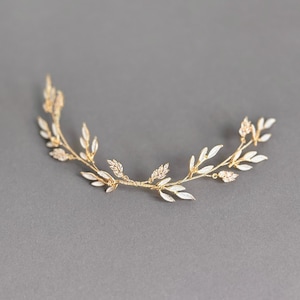 May include: A delicate gold-toned hair accessory featuring a curved design with white enamel leaves and small crystal accents. The piece is designed to be worn in the hair, likely for a special occasion.