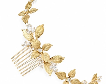 May include: A gold-toned hair comb featuring a curved, vine-like design adorned with textured metal leaves and clear marquise-cut crystals. The piece extends from a multi-pronged metal comb base, showcasing a botanical arrangement of metallic foliage.