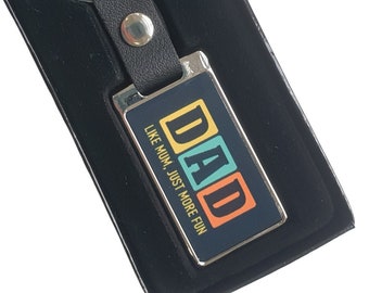 Op de afbeelding: Een zilveren sleutelhanger met een zwart leren bandje en een rechthoekig metalen label. Het label heeft een kleurrijk ontwerp met de tekst "DAD" en "LIKE MUM, JUST MORE FUN".