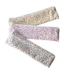 Può includere: Tre astucci rettangolari in tessuto con cerniere bianche, ognuno caratterizzato da un motivo floreale distinto. Un astuccio ha fiori gialli, uno fiori rosa e uno fiori blu, tutti su uno sfondo di colore chiaro e neutro.