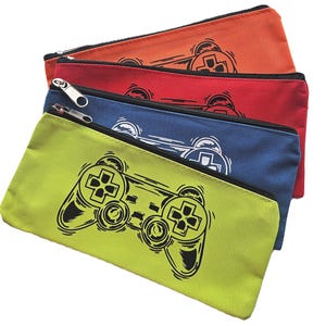 May include: Four colorful pencil cases in orange, red, blue, and green. Each case features a black and white graphic of a video game controller. The cases have black zippers and are made of a canvas-like material.
