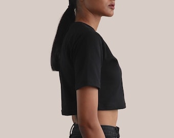Könnte beinhalten: Ein schwarzes, bauchfreies T-Shirt mit kurzen Ärmeln. Das T-Shirt wird von einer Person mit schwarzen Haaren getragen und zu einer schwarzen Jeans kombiniert.