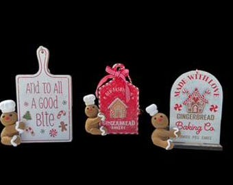 May include: A festive arrangement featuring gingerbread-themed decorations. Includes a cutting board with text, a red gift tag, and a sign, each with gingerbread men and holiday accents. The text reads "And to All A Good Bite" and "Made With Love".