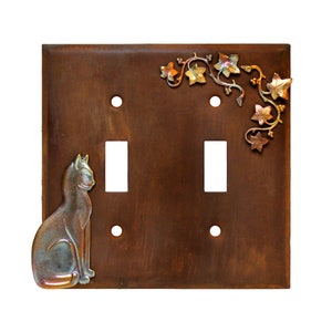 May include: A double light switch cover with a rustic brown finish. The cover features a silver cat figurine and a decorative ivy design in gold and copper.