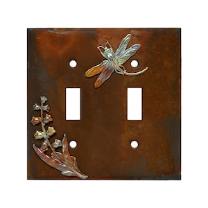 May include: A double light switch cover with a rustic brown metal finish. The cover features a dragonfly and lily of the valley design in copper, silver, and gold.