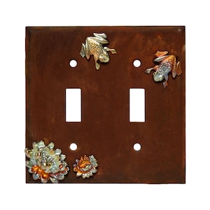 May include: A double light switch cover with a rusty brown finish. The cover features two frogs and two flower designs in gold, copper, and silver.