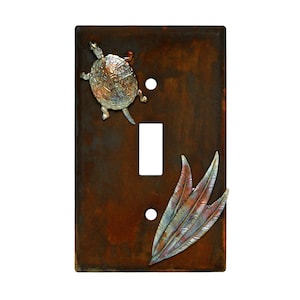 May include: A rustic brown metal light switch cover with a silver turtle and a copper and silver feather design.