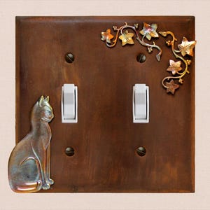Può includere: Una placca rettangolare in metallo marrone per doppio interruttore, caratterizzata da una figura di gatto in rilievo sul lato sinistro e da un ramo di edera decorativo in metallo con foglie nell'angolo in alto a destra. Due interruttori bianchi.