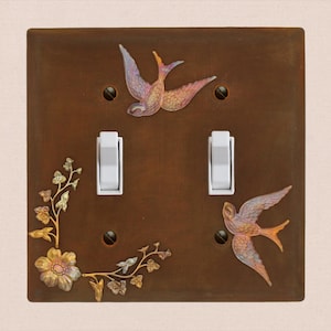 May include: A square brown double light switch plate featuring two white toggle switches. The plate is decorated with two iridescent swallows in flight and a metallic floral vine accent on the left side, secured by four dark screws.