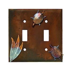 May include: A double light switch cover with a rustic brown finish. The cover features two turtles and a stylized feather design in copper and silver tones.