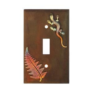 May include: A brown metal light switch cover with a fern and a lizard design. The fern is a reddish-orange color and the lizard is a blue and green color.