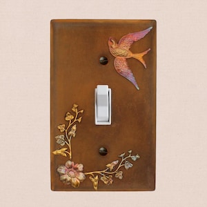 May include: A rectangular brown light switch plate featuring a white toggle switch in the center. The plate is decorated with a raised, iridescent bird in the upper right corner and a floral vine motif in the lower left corner.