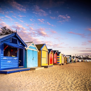 May include: A row of colorful beach huts in various colors, including blue, yellow, red, and green, line a sandy beach. The sky is a mix of blue and pink hues, with a few clouds. The huts are a popular tourist attraction.