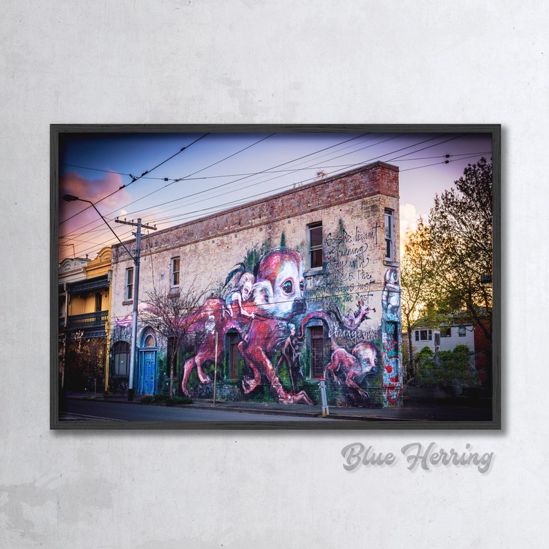 Puede incluir: Un mural colorido en un edificio de ladrillo representa una gran criatura roja parecida a un pulpo con una cara humana. El mural est&aacute; situado en el fondo de una calle de la ciudad con l&iacute;neas el&eacute;ctricas y &aacute;rboles.
