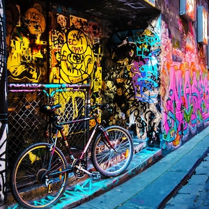 May include: A black bicycle is parked in a narrow alleyway with colorful graffiti on the walls. The bicycle has a black frame, black handlebars, and black wheels. The graffiti is in various colors, including pink, blue, green, and yellow.