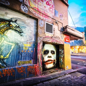 May include: A colorful street scene with graffiti art on a brick wall. The art features a skeletal creature and a portrait of the Joker. The wall is in the foreground, and a modern building with a glass facade is in the background.