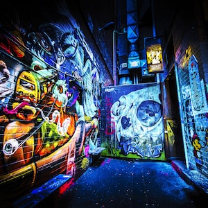 May include: A narrow alleyway with colorful graffiti art on the walls. The art features a cartoon character with a yellow helmet and a skull with a blue and white design. The alleyway is lit with blue and purple lights.