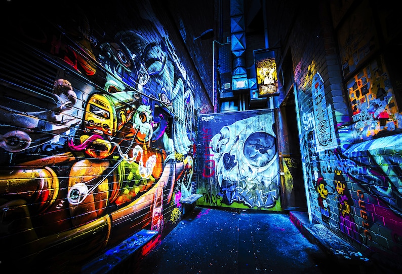 May include: A narrow alleyway with colorful graffiti art on the walls. The art features a cartoon character with a yellow helmet and a skull with a blue and white design. The alleyway is lit with blue and purple lights.