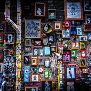 Op de afbeelding: Een bakstenen muur bedekt met ingelijste kunstwerken, waaronder abstracte schilderijen, portretten en illustraties. De kunstwerken zijn in verschillende maten en stijlen, en sommige frames zijn in felle kleuren geschilderd. De muur is ook versierd met stickers en andere ephemera.