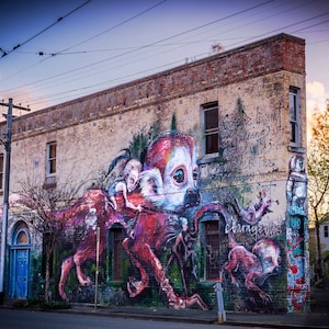 Puede incluir: Un edificio de ladrillo con un gran mural pintado en el lateral. El mural presenta una criatura roja, blanca y negra con una cara humana. La criatura sostiene una figura humana m&aacute;s peque&ntilde;a en sus brazos. El mural tambi&eacute;n incluye texto que dice "Ella no est&aacute; huyendo. Esta es la parte m&aacute;s peligrosa."