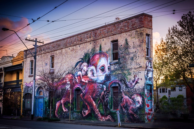 Puede incluir: Un edificio de ladrillo con un gran mural pintado en el lateral. El mural presenta una criatura roja, blanca y negra con una cara humana. La criatura sostiene una figura humana m&aacute;s peque&ntilde;a en sus brazos. El mural tambi&eacute;n incluye texto que dice "Ella no est&aacute; huyendo. Esta es la parte m&aacute;s peligrosa."