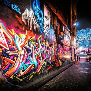 May include: A narrow alleyway in a city at night, with colorful graffiti on the walls. The graffiti features abstract shapes and designs, including a skull with a human face. The alleyway is lit by streetlights, and there are people walking in the distance.