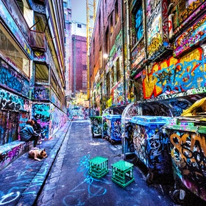May include: A narrow alleyway with graffiti-covered walls and a row of blue and green recycling bins. The bins are covered in colorful spray paint and have a variety of designs.