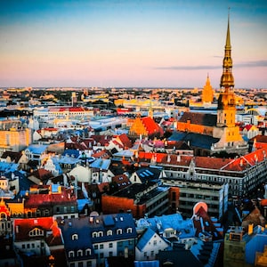 Op de afbeelding: Een luchtfoto van een skyline van een stad met een hoge, gouden torenspitskerk in het midden. De stad is gevuld met kleurrijke gebouwen en rode pannendaken. De lucht is een levendig blauw met roze en oranje tinten.