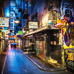 May include: A narrow, illuminated alleyway at night, lined with shops and restaurants. Graffiti art covers the walls, and signs hang overhead. The alleyway is wet, reflecting the lights, and blurred figures walk in the distance.