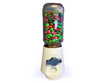 Op de afbeelding: Een witte kauwgomballenautomaat met een doorzichtige glazen pot gevuld met rode en groene snoepjes. De machine heeft een donkergrijze doseerhendel en een kleine opening aan de onderkant voor het ophalen van snoep. Op een witte achtergrond.
