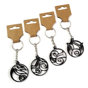 Peut inclure: Quatre porte-clés noirs avec des motifs circulaires représentant des éléments. Chaque porte-clés est attaché à une chaîne et un anneau. Ils sont présentés sur des étiquettes en carton marron, prêts à être exposés ou offerts.