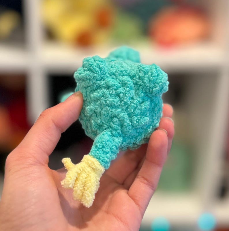 May include: A teal blue crocheted plush toy with a yellow crocheted arm. The toy is being held by a hand.