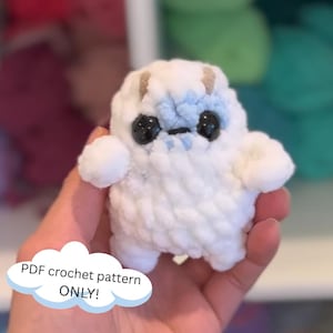 May include: A white crocheted plush toy with blue details and large black eyes. The toy is being held in a hand. PDF crochet pattern ONLY! is written on a blue cloud.