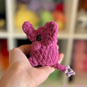 May include: A small, crocheted plush toy shaped like a purple elephant with a black button eye. The toy is held in a person's hand.