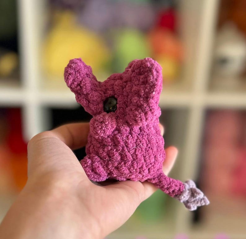 May include: A small, crocheted plush toy shaped like a purple elephant with a black button eye. The toy is held in a person's hand.