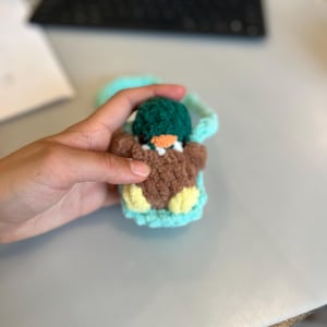 Op de afbeelding: Een handgemaakte gehaakte eend in een tealkleurige draagzak. De eend heeft een bruin lichaam, gele voeten en een groene hoed. De draagzak heeft een tealkleurige band. De snavel van de eend is oranje.