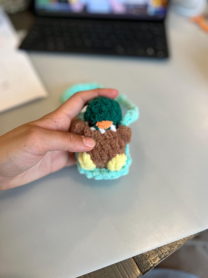 Op de afbeelding: Een handgemaakte gehaakte eend in een tealkleurige draagzak. De eend heeft een bruin lichaam, gele voeten en een groene hoed. De draagzak heeft een tealkleurige band. De snavel van de eend is oranje.