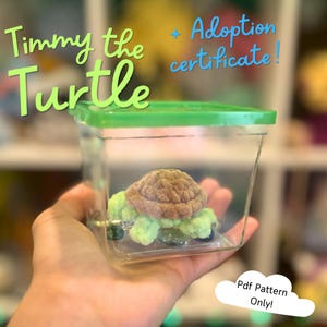Puede incluir: Un pequeño recipiente de plástico transparente con tapa verde contiene un peluche de tortuga. La tortuga tiene un caparazón marrón y un cuerpo verde. El texto en la imagen dice "Timmy the Turtle" y "Adoption certificate!" con una nube que dice "Pdf Pattern Only!"