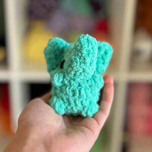 May include: A small, crocheted elephant toy in a bright turquoise color. The elephant has a textured surface and is held in a person's hand.