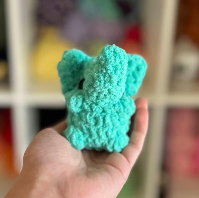 May include: A small, crocheted elephant toy in a bright turquoise color. The elephant has a textured surface and is held in a person's hand.