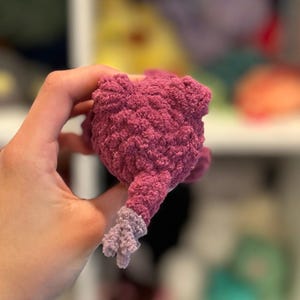 May include: A plush purple and lavender crocheted toy with a textured surface. The toy is shaped like a small animal with a tail.